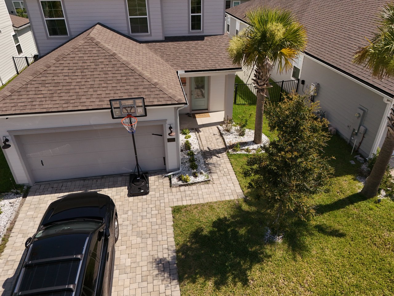 Aerial view of a home’s entrance and garage, with a paver driveway, basketball hoop, and palm tree.