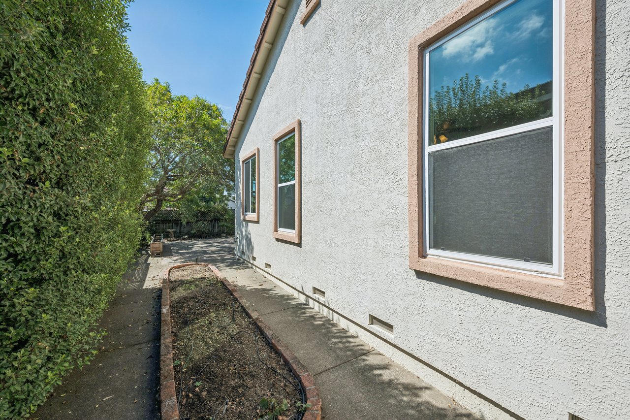 House side with tan-trimmed windows beside a narrow walkway and hedge-lined planting bed.