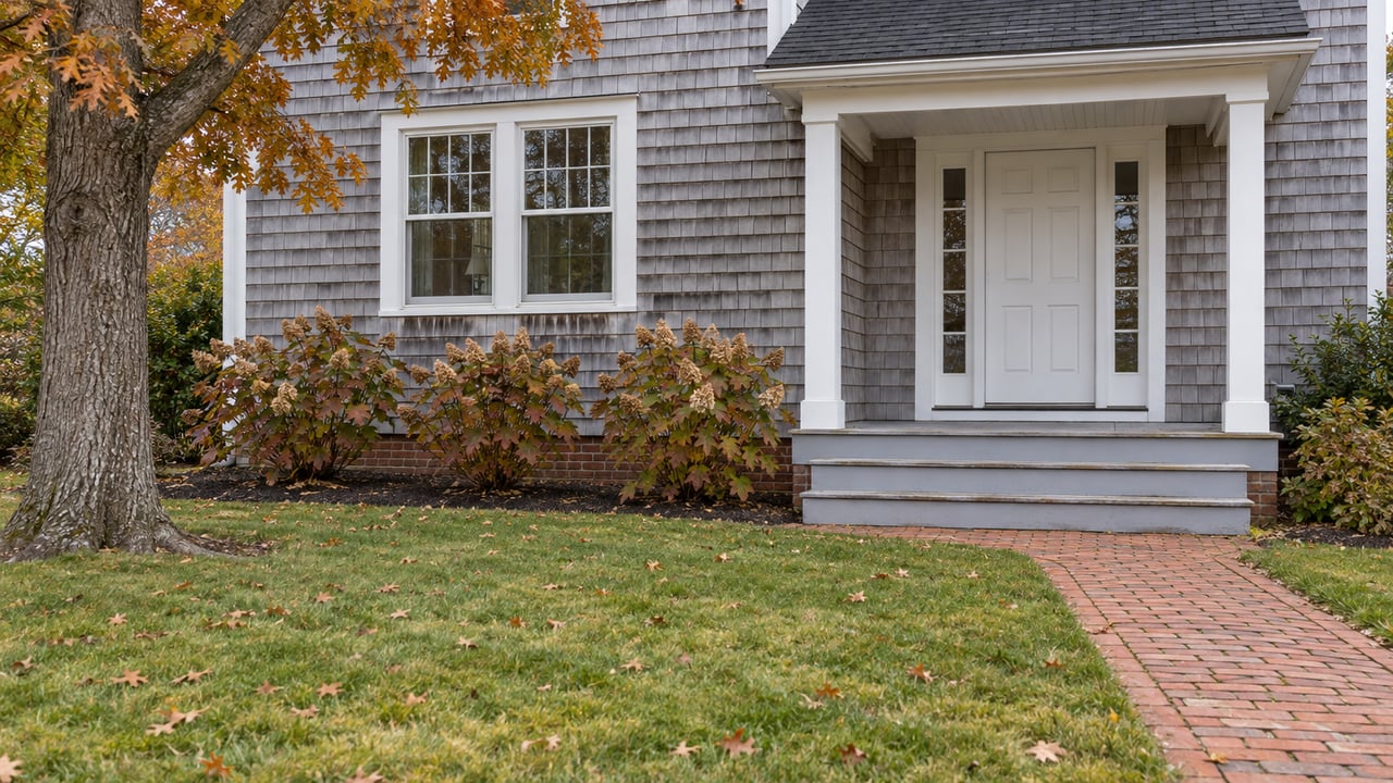 Cedar-shingled home with a covered white-trimmed entrance, brick walk, autumn hydrangeas, and a deep green front lawn.