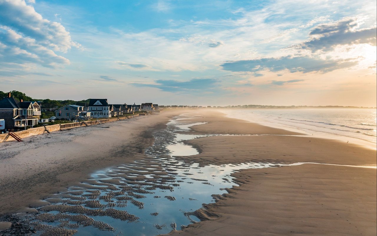Coastal beach at low tide with oceanfront houses and reflective tidal pools in warm evening light.