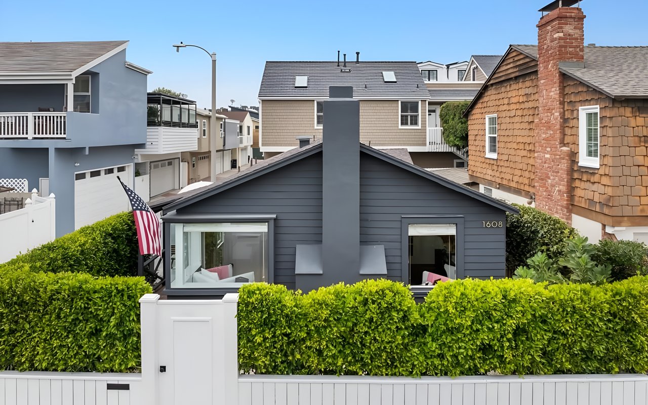  A modern gray home with a white picket fence, a tall hedge, and an American flag flying by the front window.