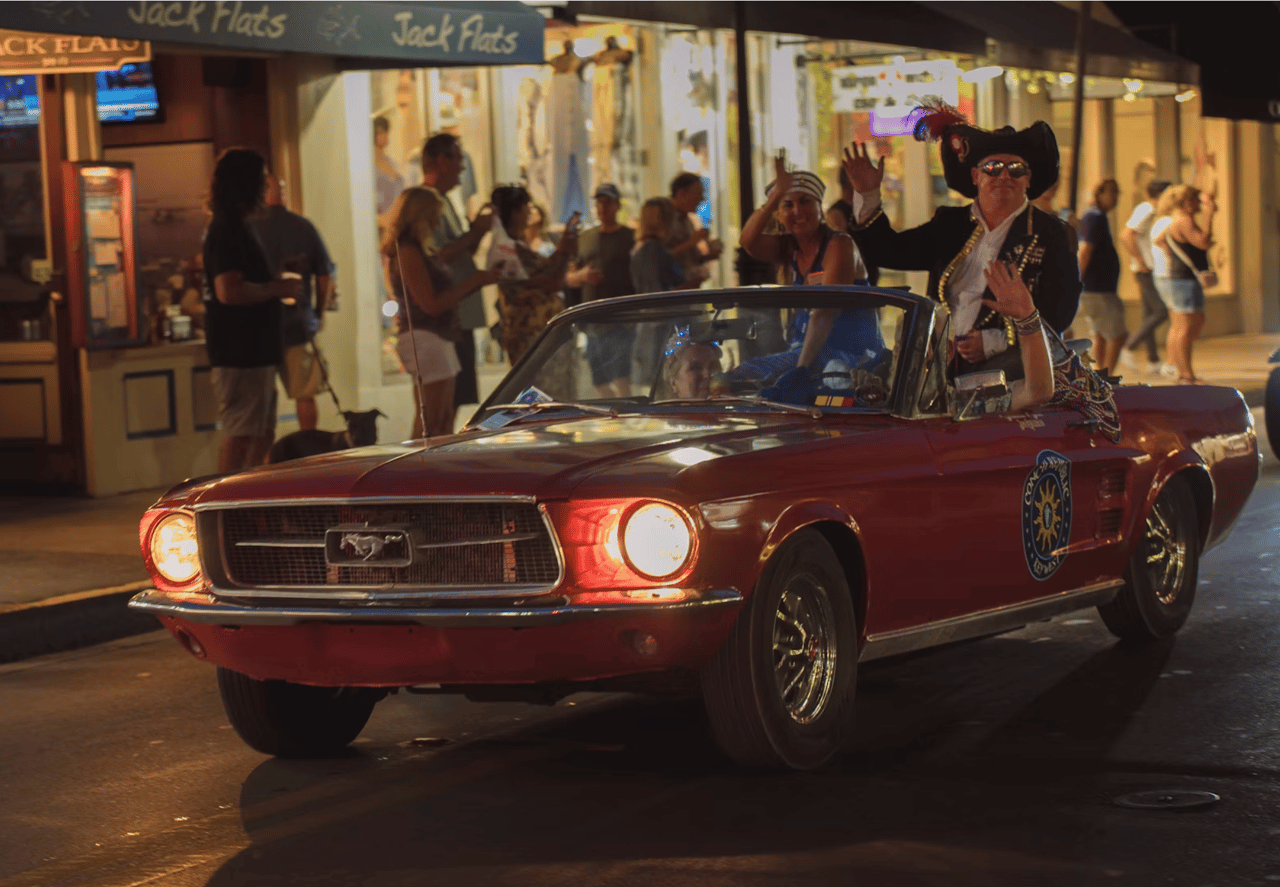 Red vintage Mustang convertible on a busy night street, with a feathered-hat passenger waving.