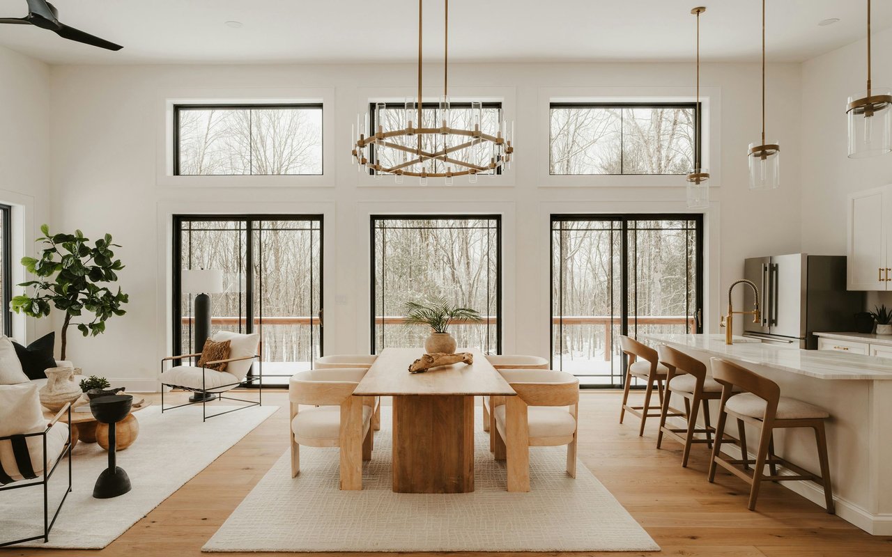 Open-plan dining and living area with a wood table and tall windows overlooking snowy woodland.