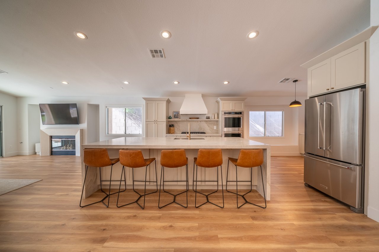 Open-plan kitchen with a stone island, five rust-colored stools, white cabinets, and stainless steel appliances.