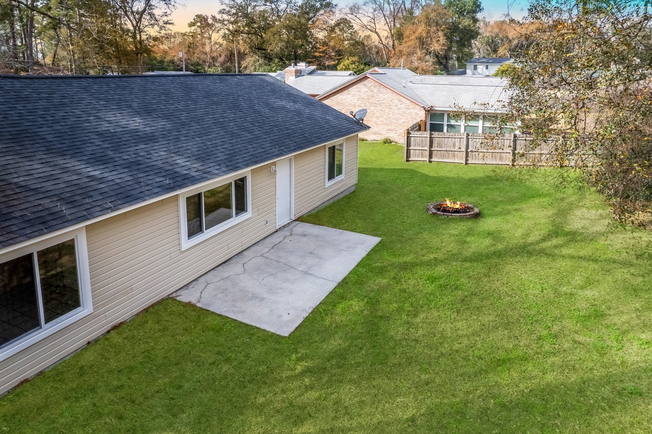 Aerial view of a backyard with a beige house, concrete patio, and grassy lawn. A small fire pit is lit, and fall trees create a calm, cozy atmosphere.