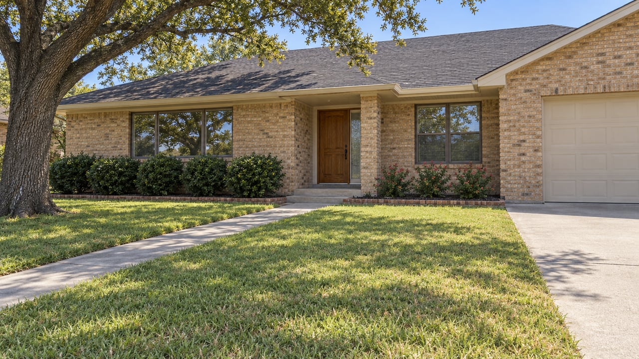 A buff-brick single-story home with a deep front lawn, broad oak canopy, and a concrete path leading to a sheltered wood entry.