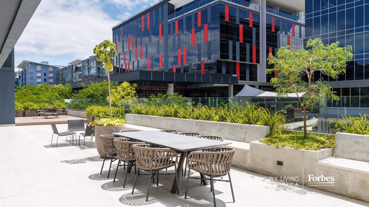 Apartment terrace with an outdoor dining set, raised planters, and modern glass buildings beyond.