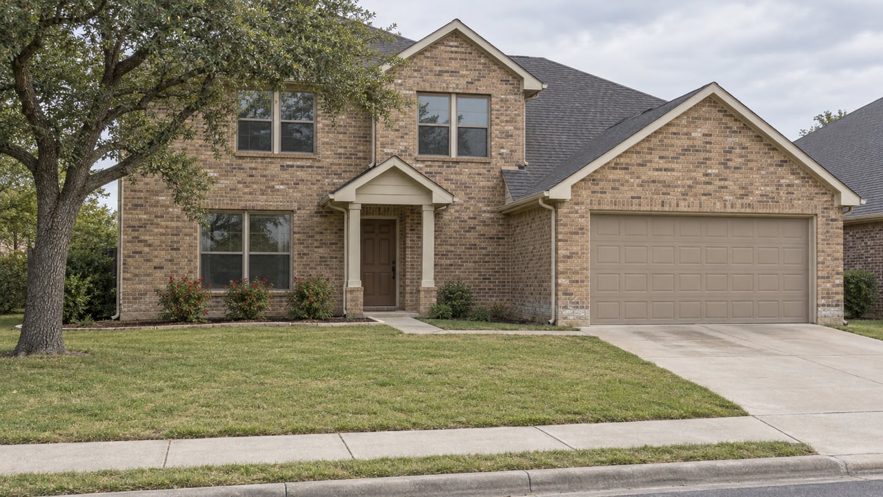 Two-story tan brick house with a front-facing garage, broad lawn, mature live oak and concrete driveway under soft daylight.