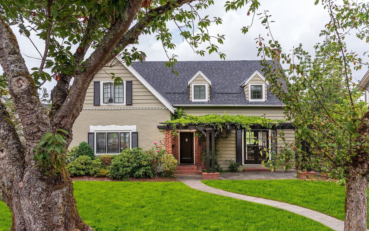 Beige two-story house with dormers and brick entry, framed by mature trees and a green lawn.