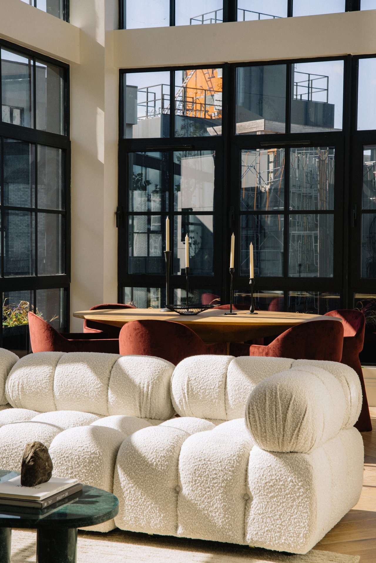 Living and dining area with a textured white sofa, rust-colored chairs, and tall black-framed windows.