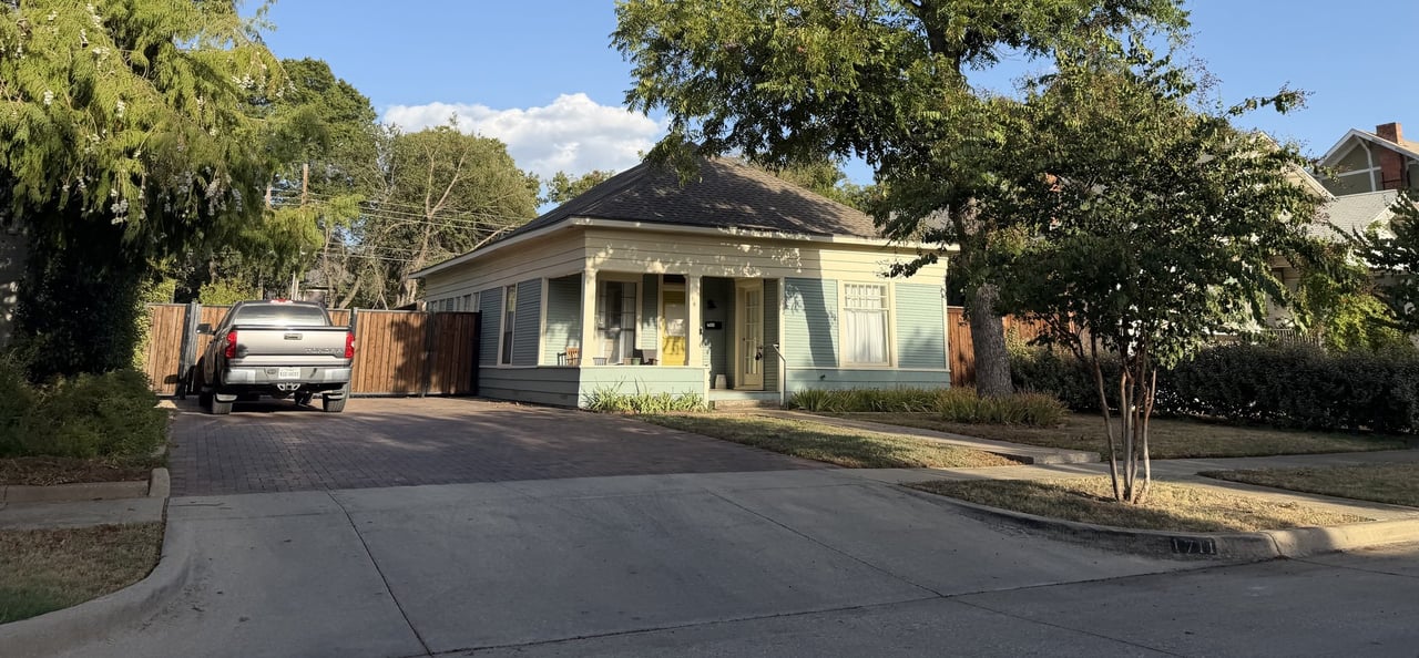 Single-story cottage with a covered porch, mature shade trees, and a paved driveway.