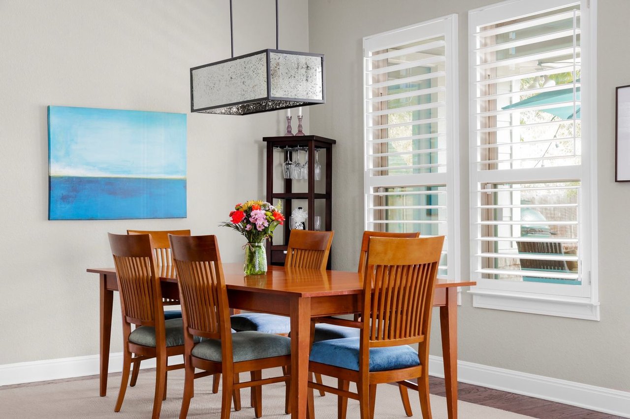 Dining room with a wooden table, colorful flowers, blue artwork, and tall shuttered windows.