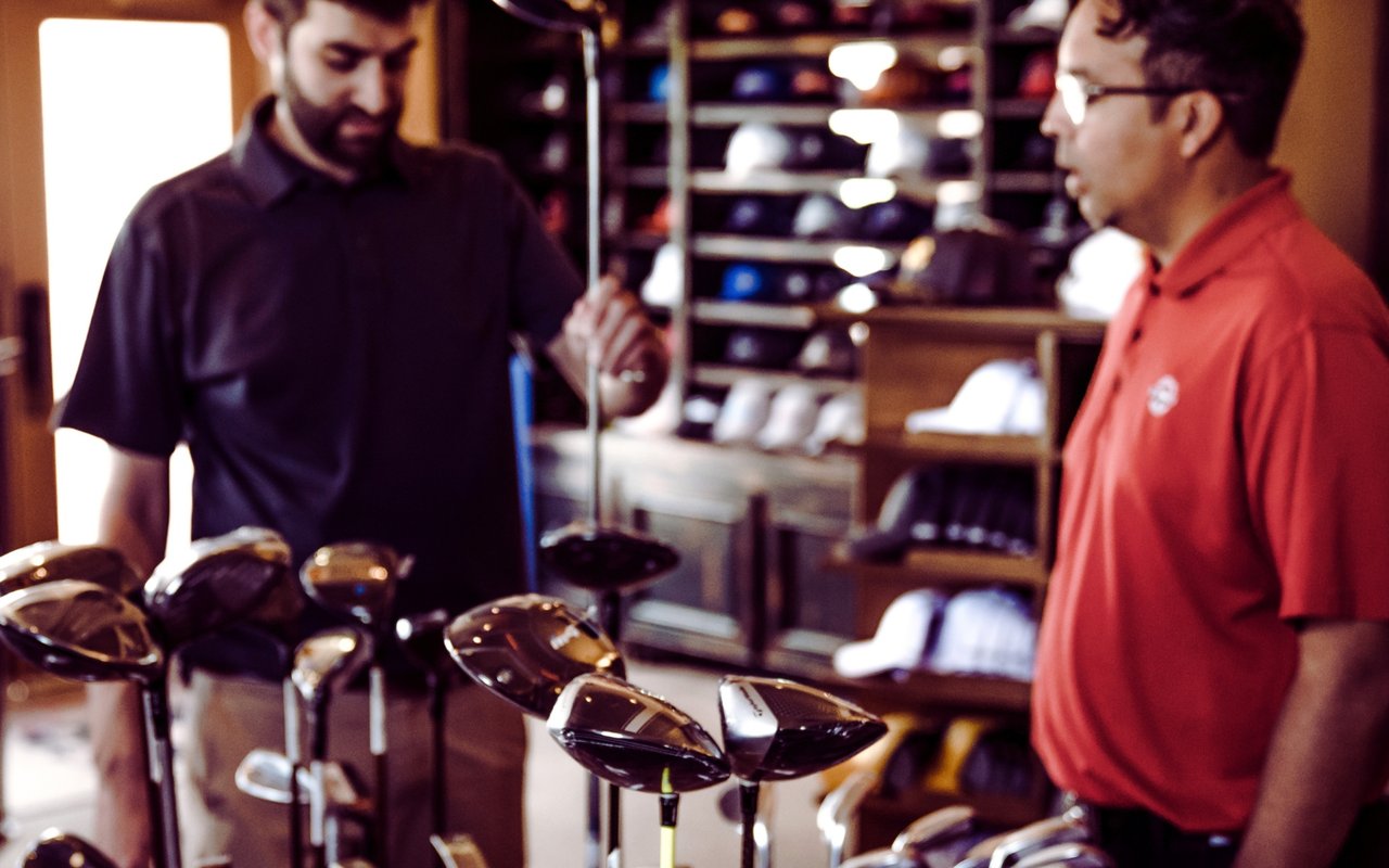 Two men discuss golf clubs in a shop, with rows of club heads displayed in front of them.