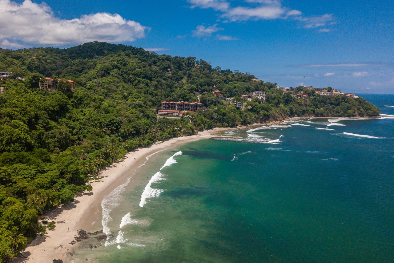 Tropical coastline with a sandy beach, turquoise water, and buildings among forested hills.