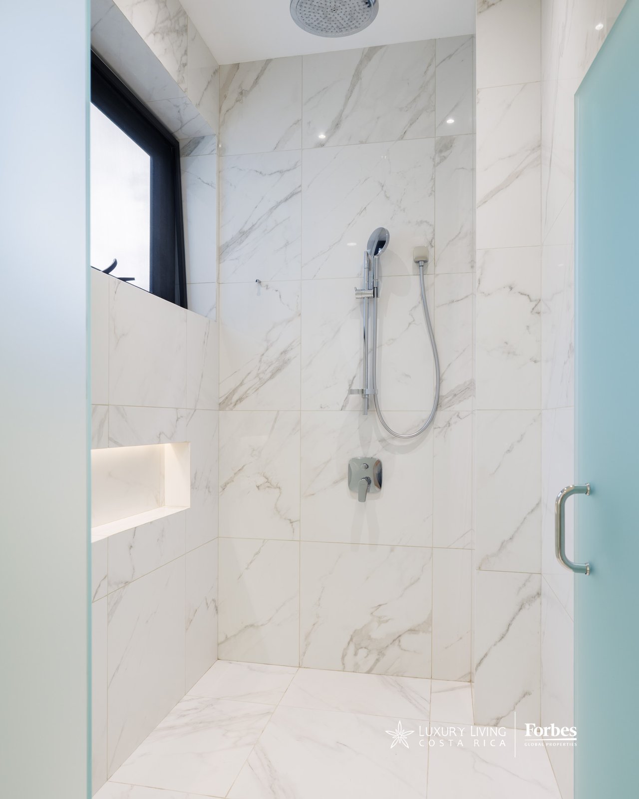 Marble-tiled walk-in shower with a rain showerhead, handheld wand, and illuminated recessed niche.