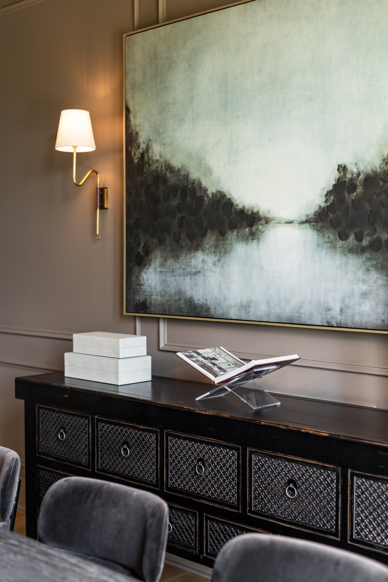 Staged interior with a carved console, upholstered chairs, abstract painting, and brass wall lamp.