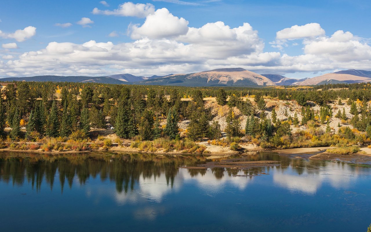 Mountain lake reflecting evergreen forest and autumn-colored slopes beneath a cloudy blue sky.