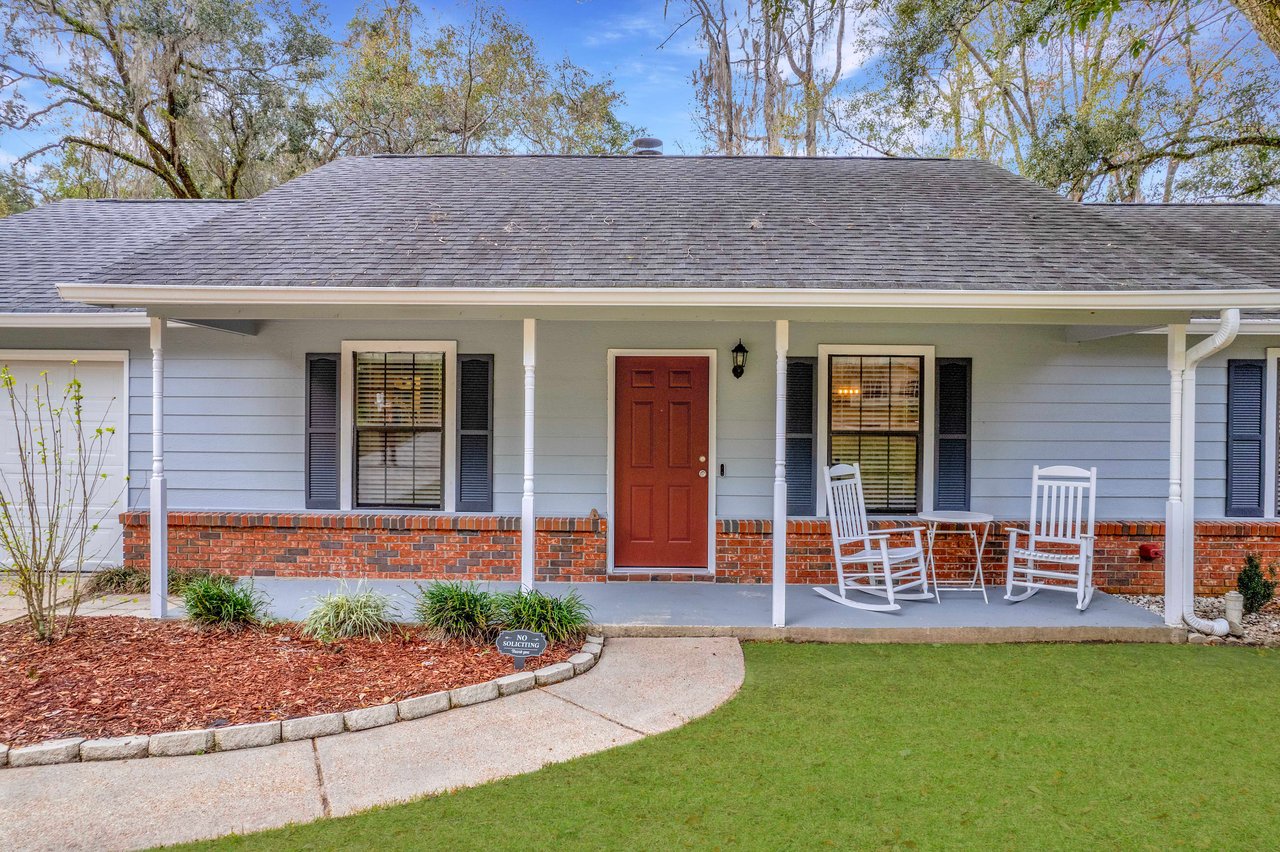 Front view of a cozy house with a light blue exterior and a red door. Two white rocking chairs sit on the porch, conveying a peaceful, welcoming tone.