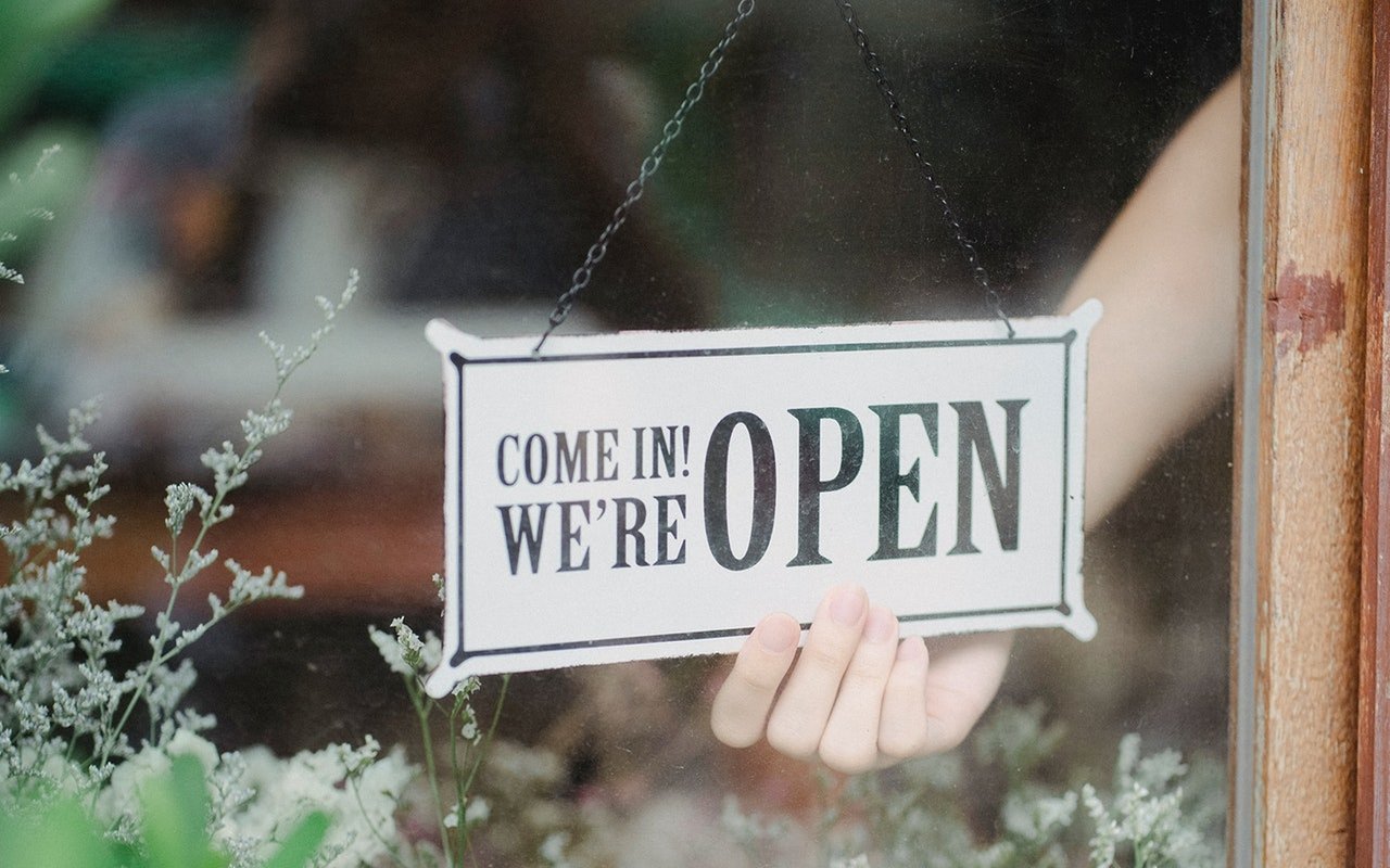 An open sign hangs in a shop window as a hand holds it from behind.