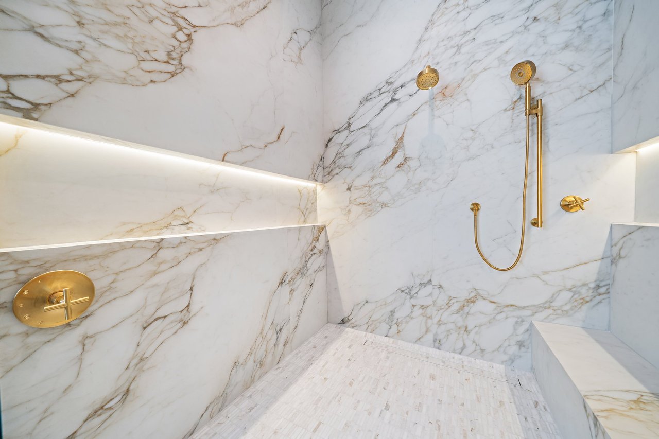 Marble walk-in shower with illuminated shelves, a bench, and gold fixtures.