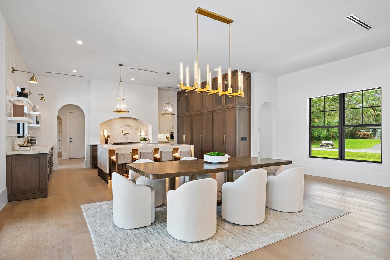 Open kitchen and dining area with dark wood cabinetry, marble backsplash, and a large table beneath brass lighting.