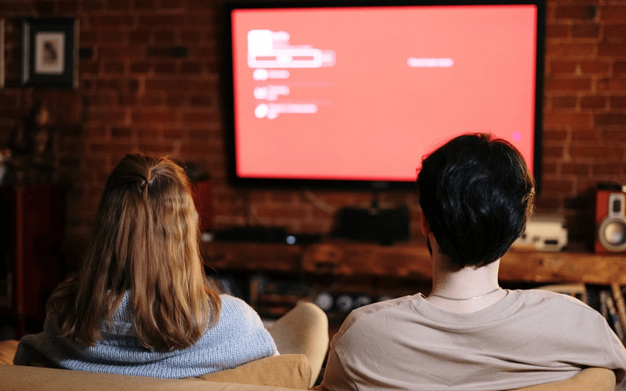 Two people watch a red-glowing television from a couch in a brick-walled living room.