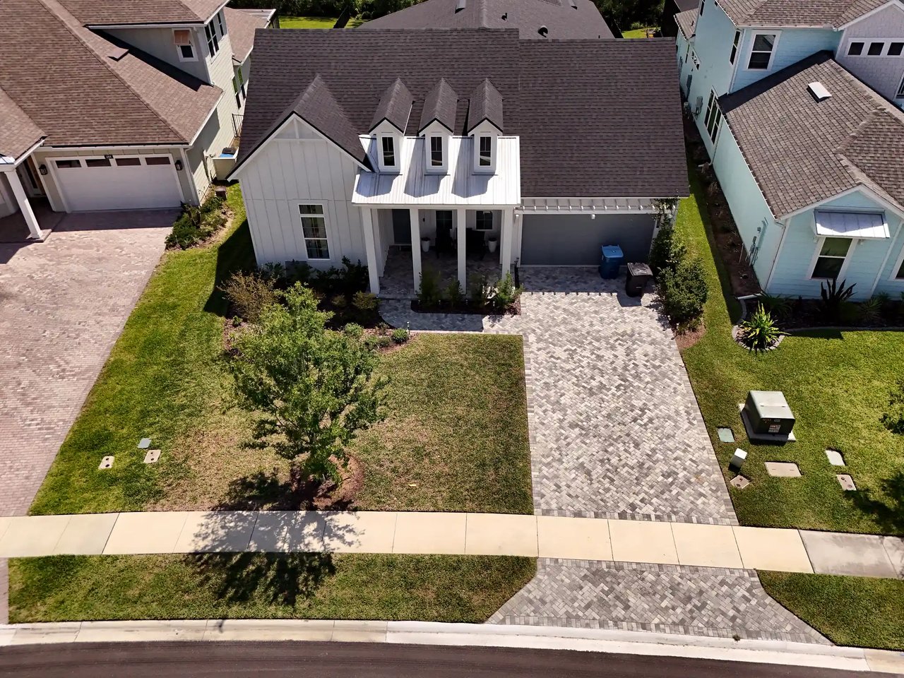 Elevated view of a white single-story farmhouse-style home with three dormers, a covered porch and a paver driveway.