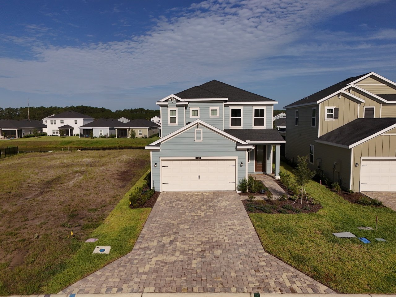 Two-story light blue home with a white double garage door and wide paver driveway in a new neighborhood.