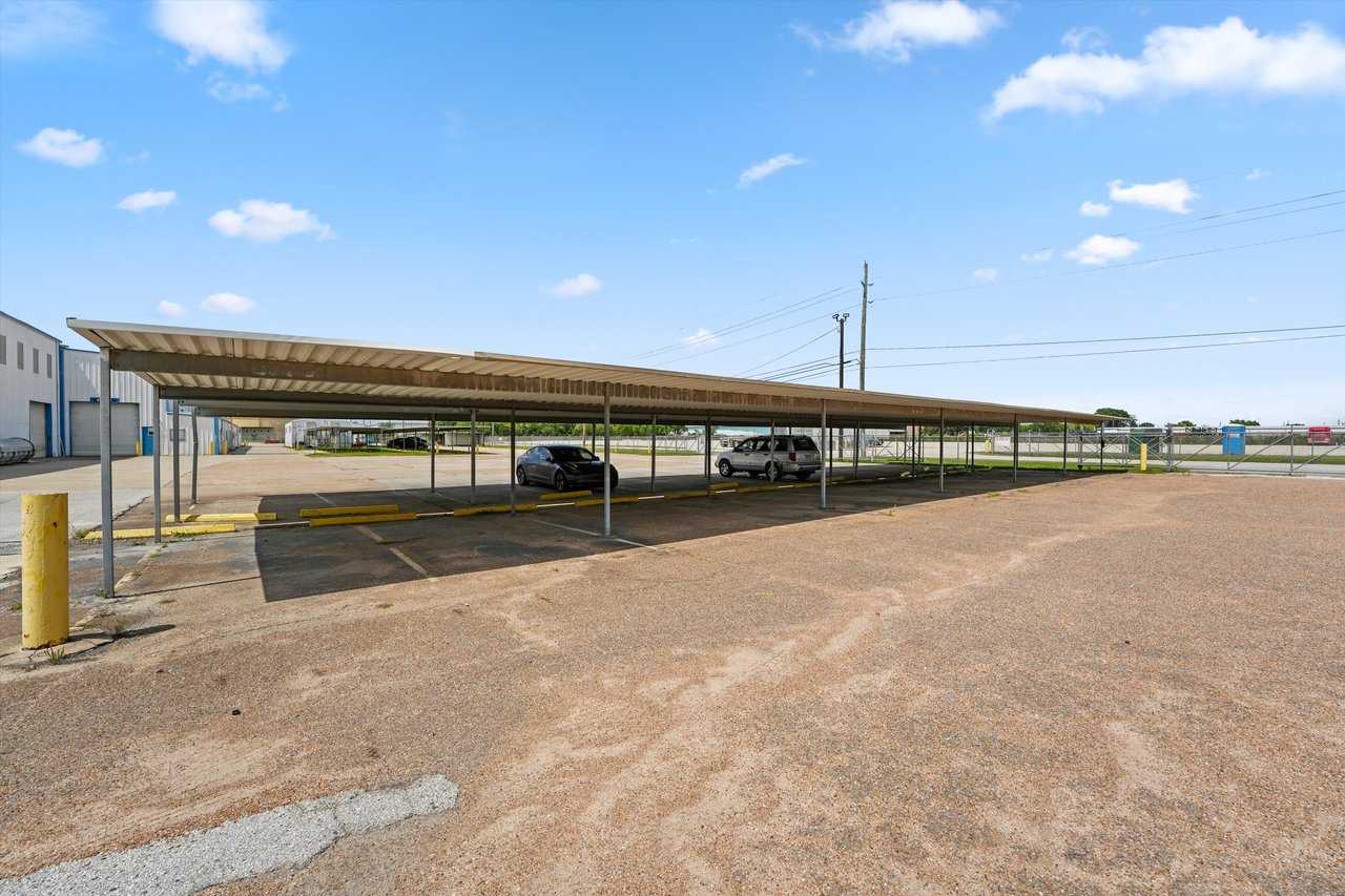 Warehouse property with a long covered parking canopy, two cars, and a paved lot.