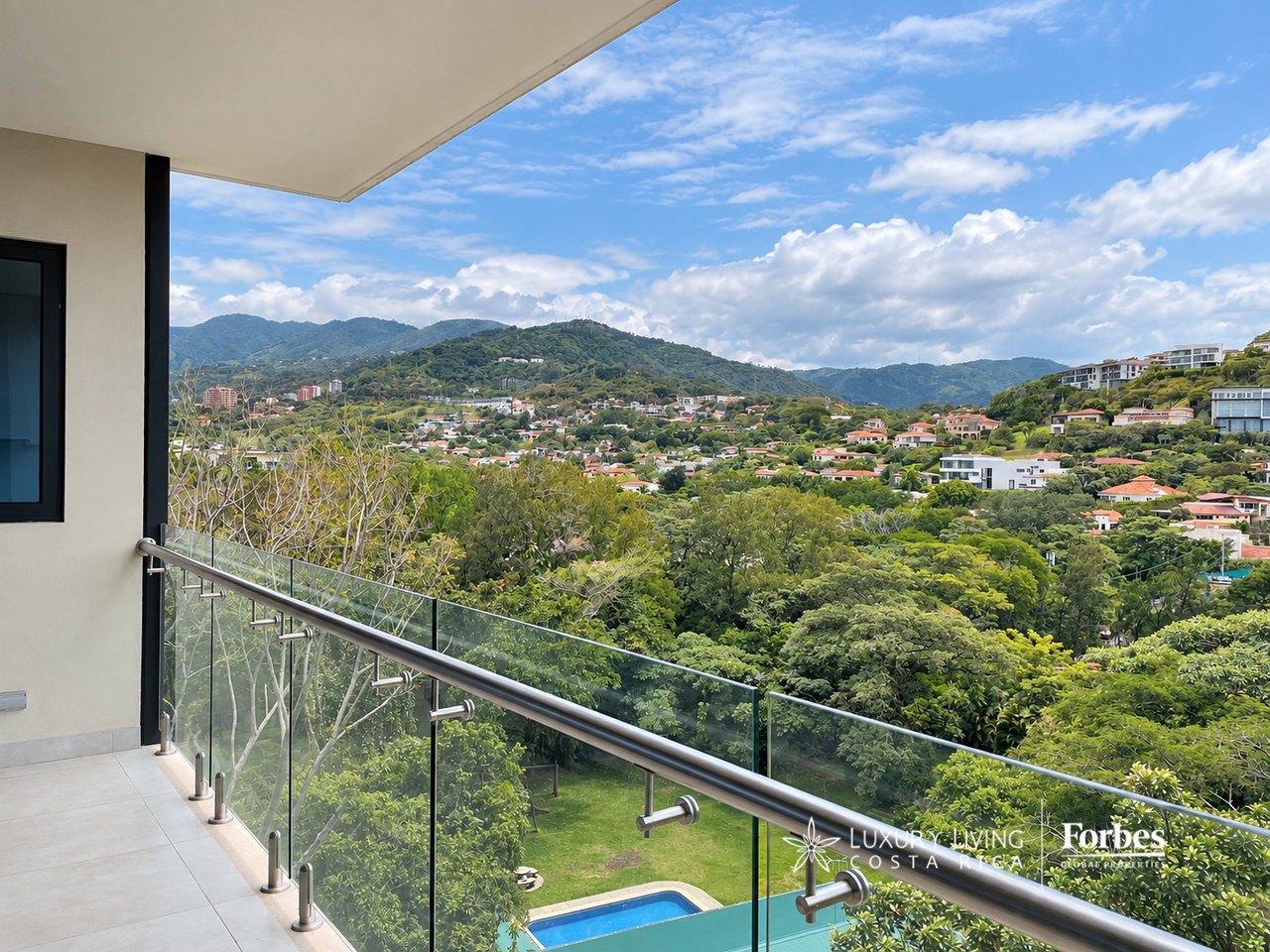 Apartment balcony with glass railing overlooking a pool, hillside homes, and green mountains.