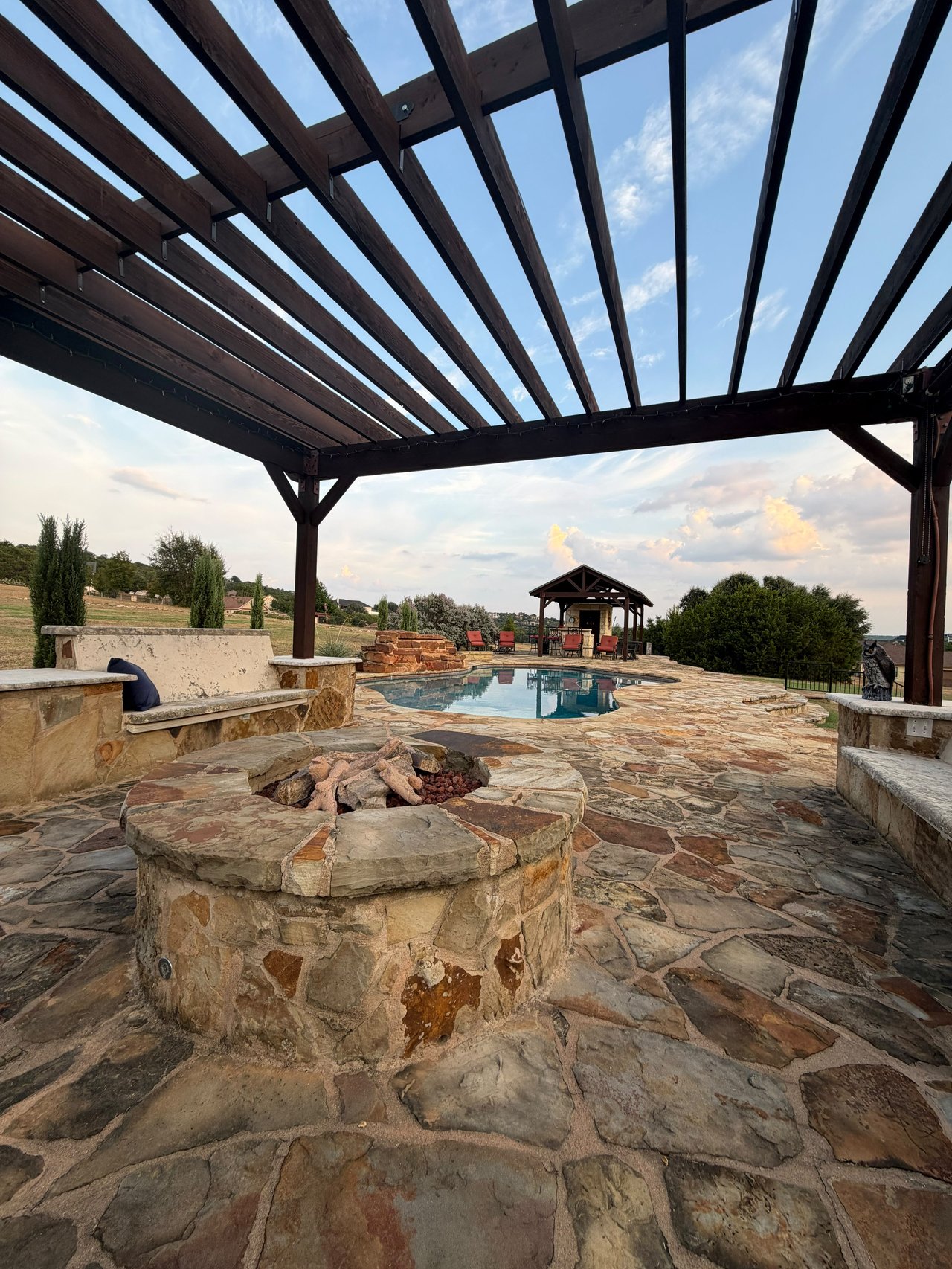 Stone pool patio with a timber pergola, fire pit, and distant covered seating.