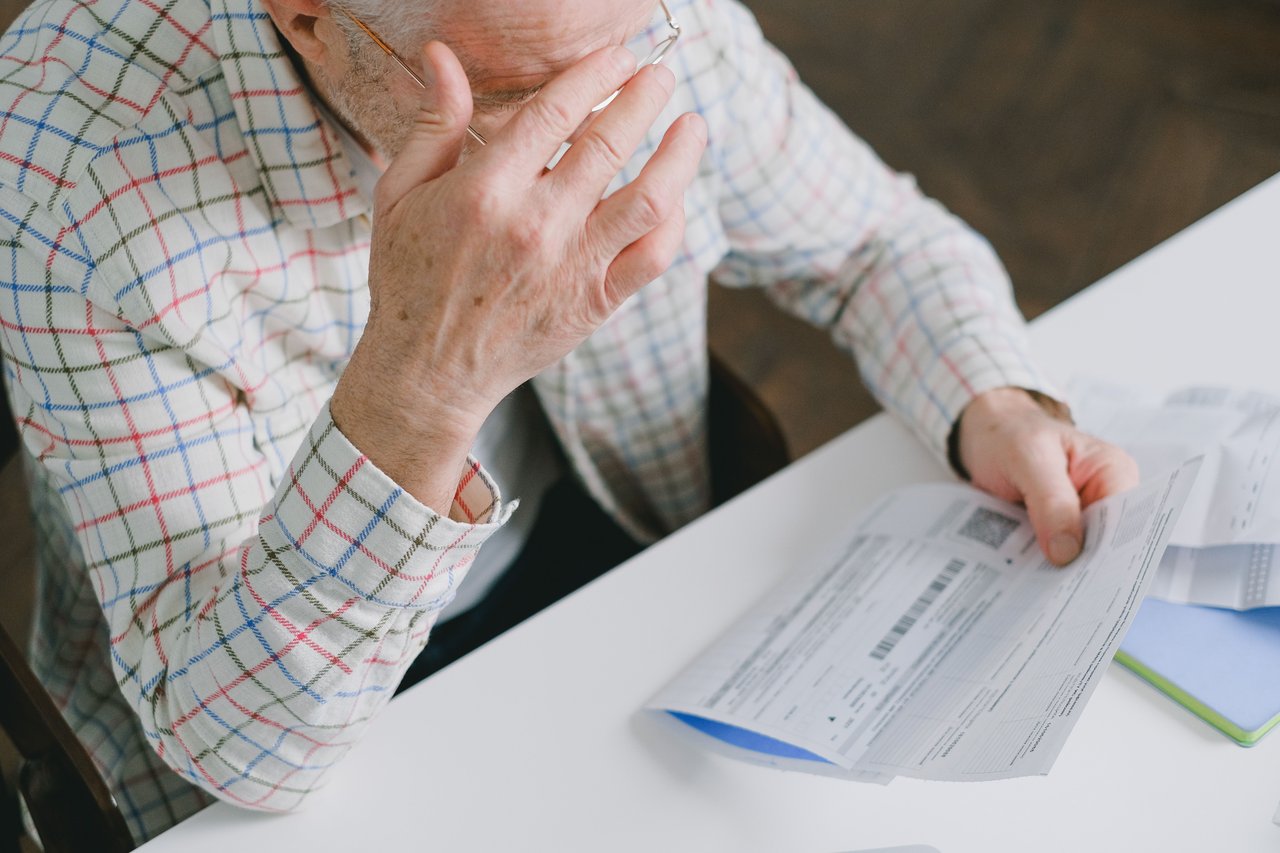 Older man in glasses rubs his face while reviewing paperwork at a table.