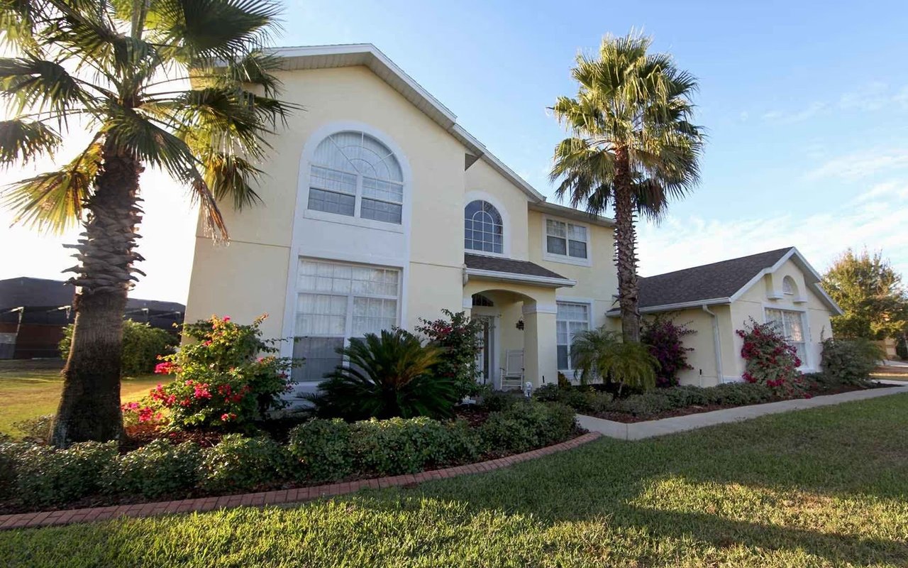 Two-story cream house with arched windows, palm trees, and flowering shrubs along the front garden.