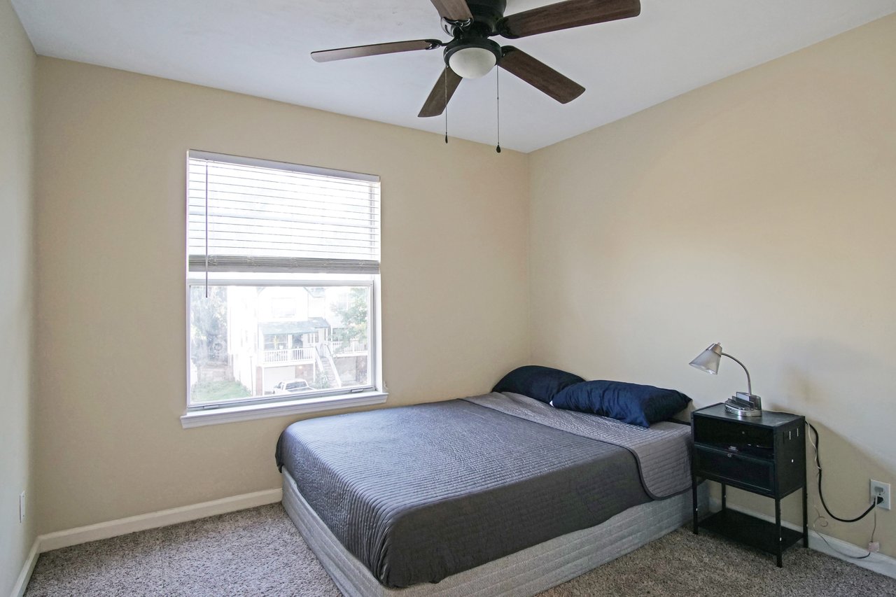 Bedroom at 529 Collinsford Road, Tallahassee, Florida, featuring a large window with blinds, neutral beige walls, carpet flooring, and a ceiling fan with light fixture. The room includes a bed with gray bedding and a black nightstand with a reading lamp.