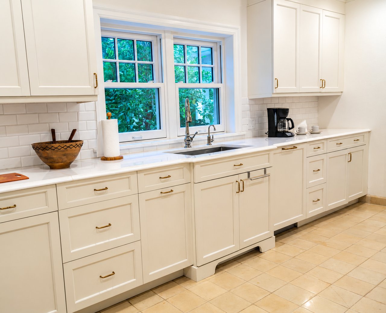White kitchen with brass cabinet pulls, a sink beneath two windows, and greenery outside.