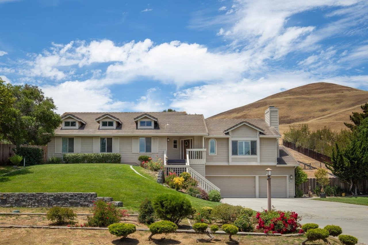 Light-colored home with dormer windows and a landscaped lawn below dry hills.