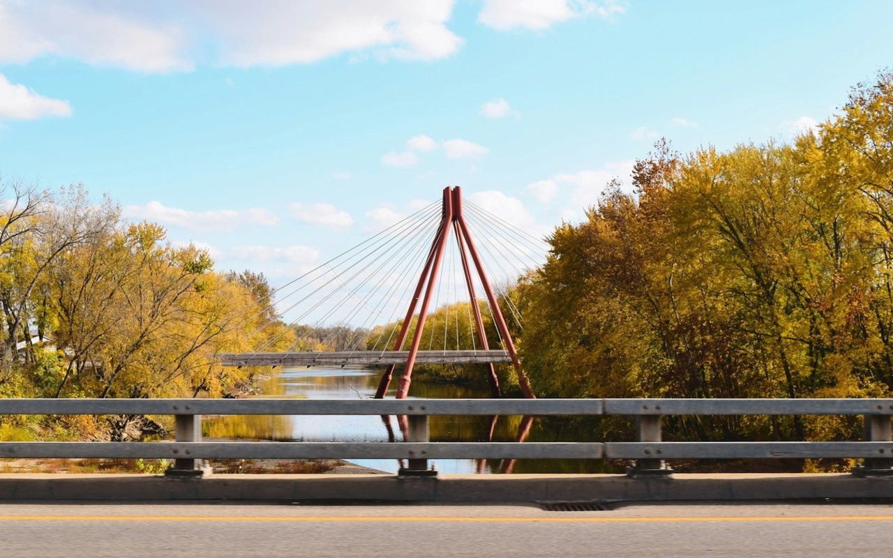 Red cable-stayed bridge over a river, framed by golden autumn trees and a road bridge.