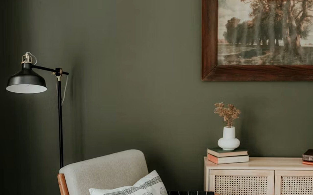 Living room corner with a cream chair, black floor lamp, and cane-front cabinet against an olive-green wall.