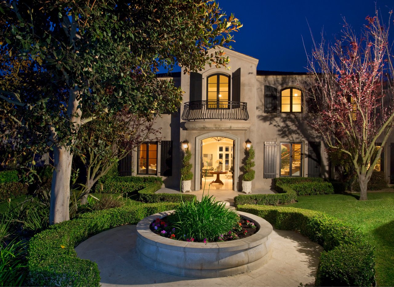 Two-story home at night with a wrought-iron balcony and circular courtyard planter.