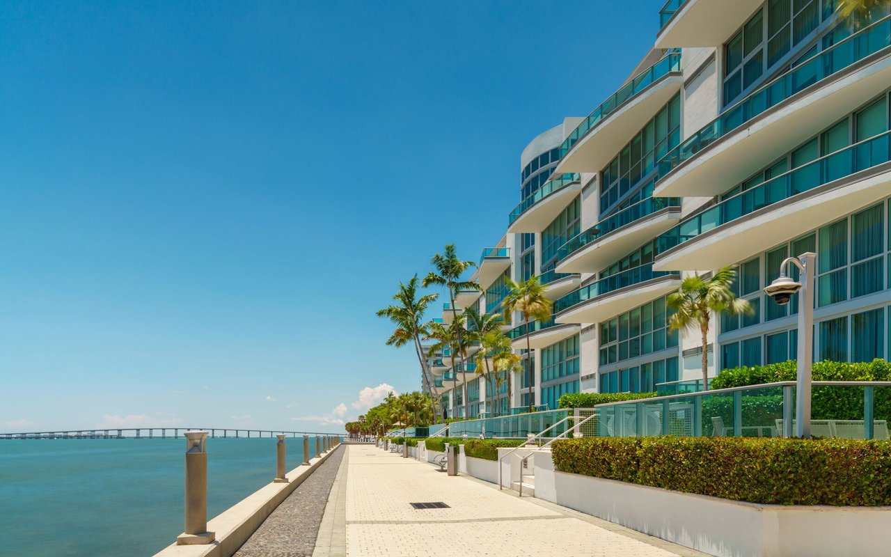 Waterfront condominium with glass balconies beside a palm-lined promenade and calm blue water.