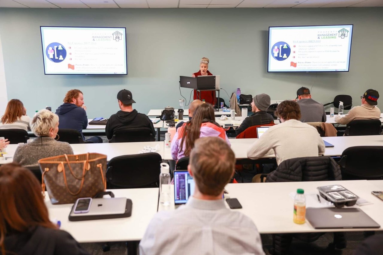 A presenter leads a real estate training session as attendees sit at classroom tables.