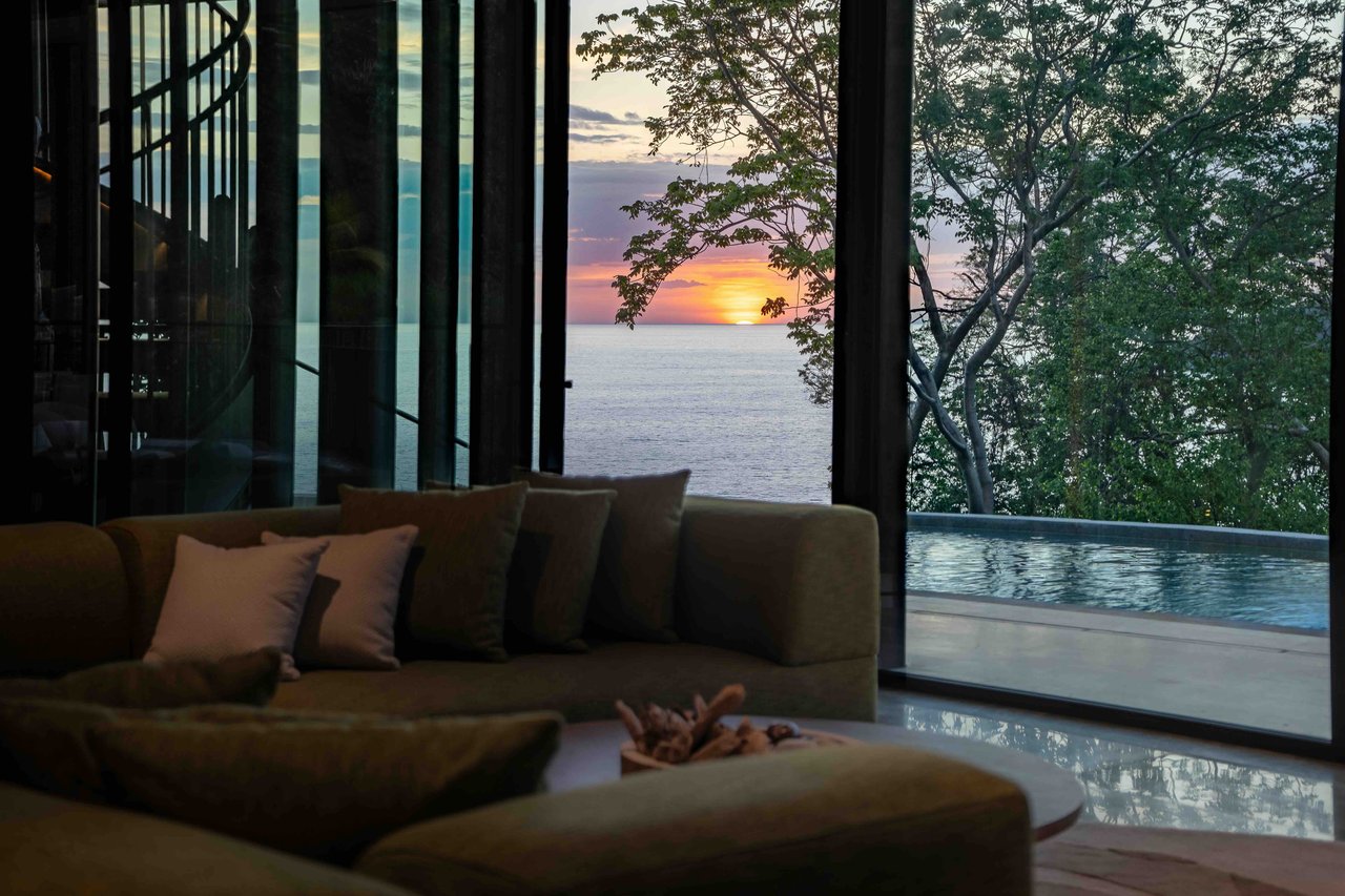 Living room with floor-to-ceiling windows overlooking an infinity pool and ocean sunset.