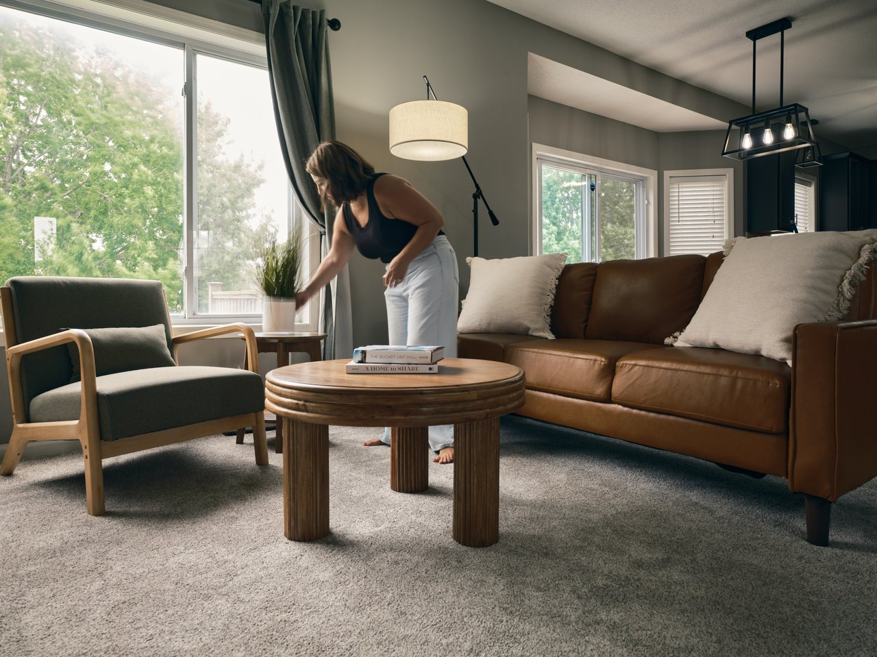 Staged living room with a brown leather sofa, green armchair and woman arranging decor by a large window.