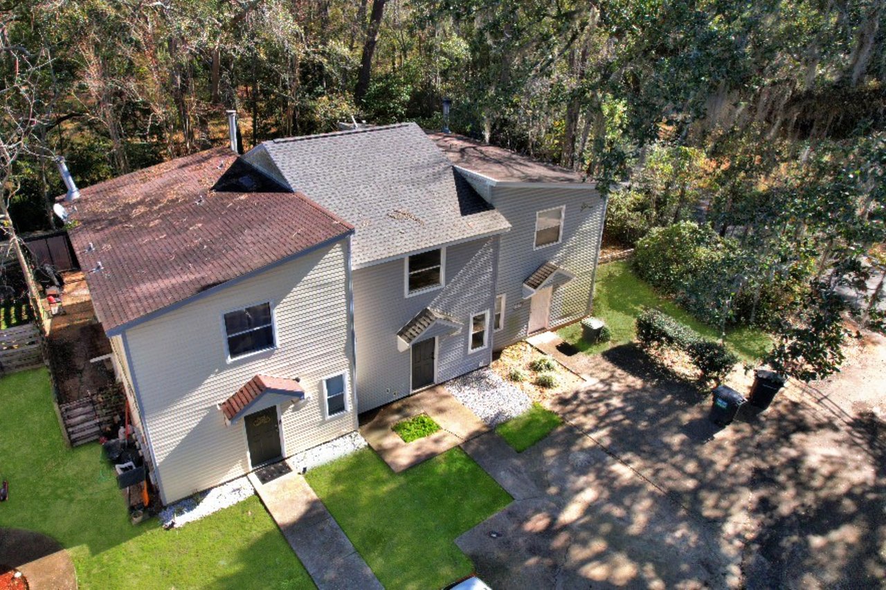 Aerial view of a beige duplex surrounded by trees. Each unit has a dark door and window awnings. Sunlight casts shadows on the vibrant green lawn.