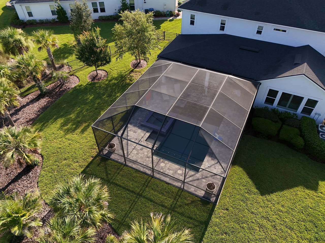 Aerial view of a white home’s screened backyard patio, landscaped lawn, and palm trees.