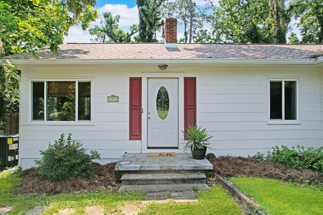Small white house with a brick chimney, red shutters, and a welcoming white door. A potted plant and a doormat saying "hello" adorn the entrance.