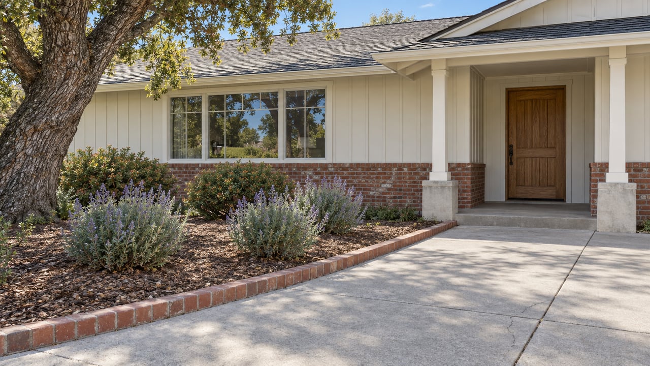 Single-story cream-sided ranch with brick accents, a broad concrete approach and sculptural oak beside drought-tolerant planting.