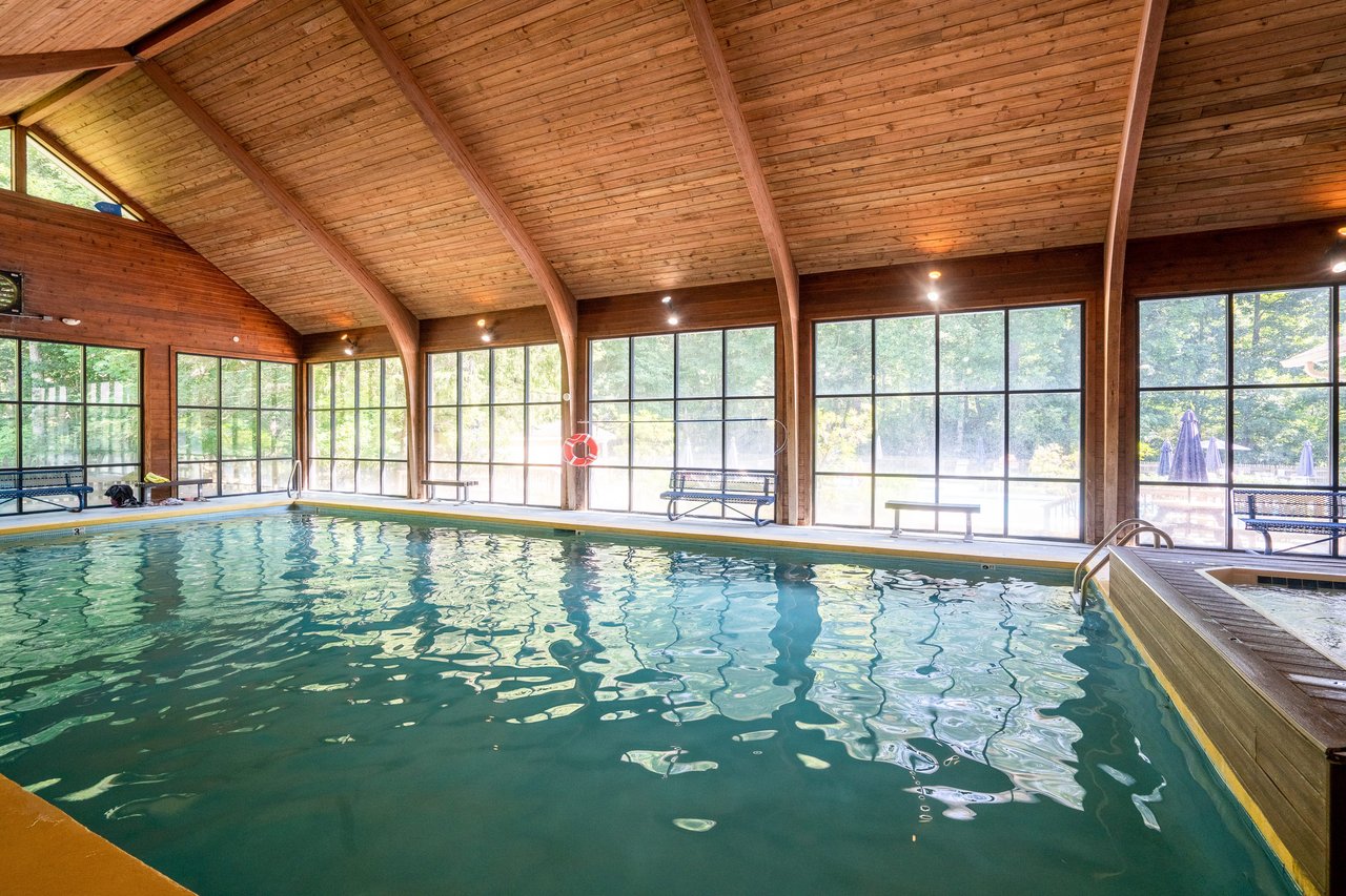 The indoor swimming pool features large windows and a wooden ceiling, with benches along the sides for relaxation. Natural light floods the space, highlighting a lifebuoy on the wall. This serene setting offers real estate agents an impressive platform to showcase potential home features.