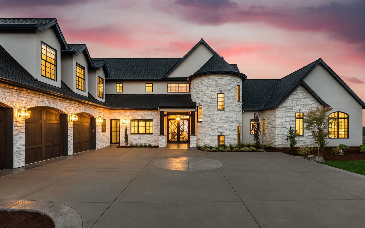  A majestic, sprawling two-story house with a mix of light-colored stone and dark trim under a dark roof, illuminated by warm interior lights at dusk.