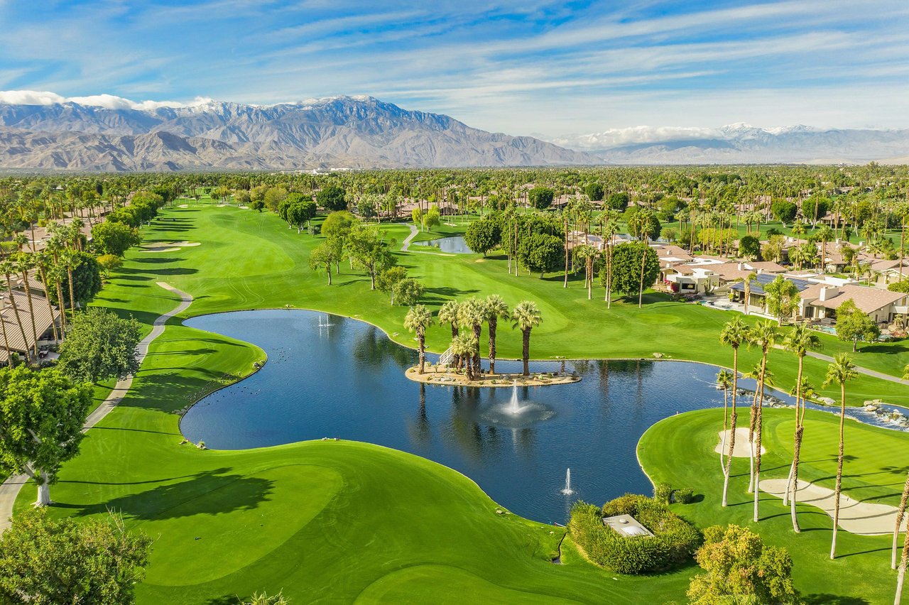 Aerial of The Lakes Country Club in Palm Desert, a lake with a palm island and fountains, fairways, and condominium rows below snowcapped mountains.